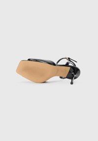 Sándal de tacón alto negro con una parte superior lisa, fina correa para el tobillo y una suela texturizada color beige. Presenta un diseño elegante con punta afilada.