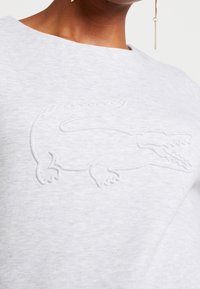 Světle šedá mikina s embosovaným logem krokodýla na hrudi. Vyrobena z měkké látky, má kulatý výstřih a minimalistický design.