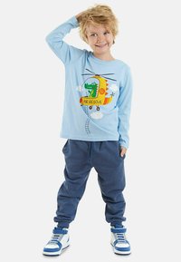 Camicia a maniche lunghe azzurro chiaro con una grafica di un elicottero e un coccodrillo, abbinata a pantaloni jogging blu scuro e sneaker bianche/blu.