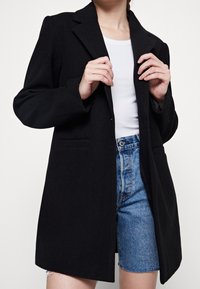 Cappotto nero su misura con collo a rever, maniche lunghe e tasche laterali, abbinato a una maglietta bianca e pantaloni short in denim. Texture liscia.
