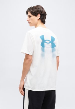 Joven mirando hacia la izquierda, vestido con una camiseta blanca con un gran logo azul de Under Armour en la parte posterior y pantalones negros con rayas blancas.