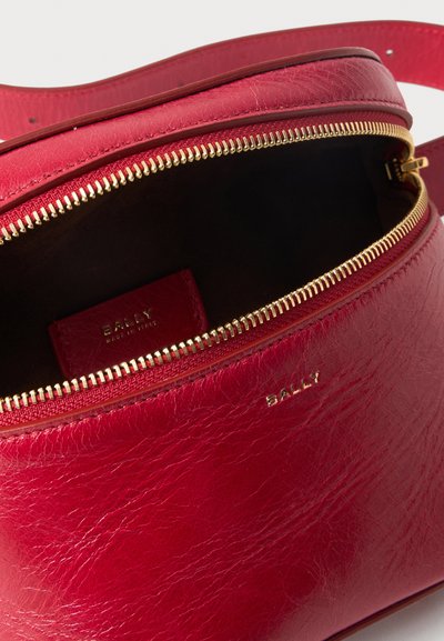 Κόκκινη δερμάτινη τσάντα μέσης Bally με χρυστό φερμουάρ μερικώς ανοιχτό, που δείχνει το σκούρο εσωτερικό και το λογότυπο Bally μπροστά και στο εσωτερικό.