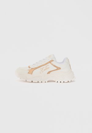 White athletic shoe with textured surface, gold accents, and a chunky sole. Features laces and brand detailing on the side.
