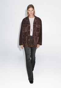 Veste en daim marron avec quatre poches, fermeture à boutons et coupe décontractée. Portée sur un haut blanc et associée à un jean évasé foncé.