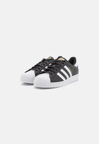 Een paar zwart-witte Adidas Superstar sneakers met shell-toes, drie witte strepen, zwarte veters en gouden branding op een witte achtergrond.