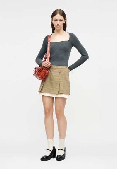 Femme portant un haut gris à manches longues, une jupe à carreaux beige, des chaussettes blanches, des talons noirs, et portant un sac à bandoulière en cuir rouge.