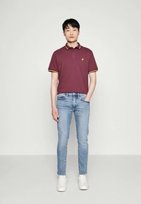 Camisa polo marrón con ribetes amarillos, combinada con jeans azul claro y zapatillas blancas; diseño simple, mangas cortas y un corte relajado.