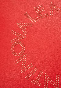 Czerwona skórzana torba z złotymi literami w formie strefy okręgowej. Tekstura jest drobno ziarnista, z zaokrąglonymi krawędziami i wyraźnym logo.