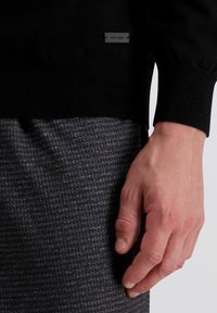 Schwarzer Strickpullover mit gerippten Bündchen und einem kleinen Markenetikett. Graue strukturierte Hose mit subtilen Mustern und lockerer Passform.