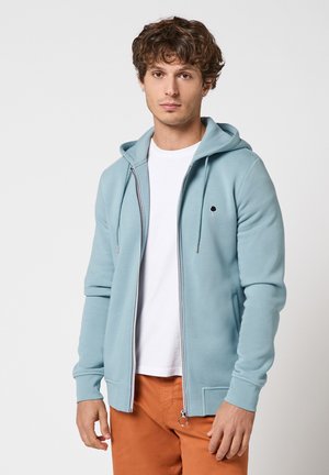 Jeune homme portant un sweat à capuche zippé bleu clair sur un t-shirt blanc et un pantalon orange, sur un fond clair uni.