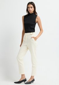 Vrouw in een mouwloos zwart topje, crèmekleurige high-waist broek met riemdetails en zwarte schoenen met lage hakken, staand tegen een effen achtergrond.