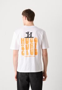 T-shirt in cotone bianco con testo grafico nero e arancione sulla parte posteriore: "HUGO SURF CLUB", con "POWERED BY WAVES" sotto. Maniche corte.