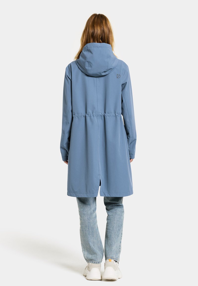 Wasserdichte blaue Parka mit Kapuze, tailliertem Schnitt und Rückenbelüftung, kombiniert mit hellblauen Jeans und weißen Turnschuhen.