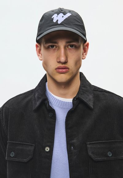 Jeune homme portant une casquette noire avec un logo abstrait blanc, une veste en velours côtelé noire et un pull bleu clair sur fond blanc uni.