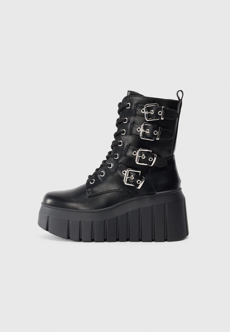 Botte de combat plateforme noire avec semelle épaisse à rainures, devant à lacets et quatre sangles à boucles argentées sur le côté.