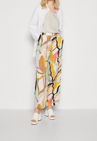 Blommönstrade vida byxor i ljusa pastellfärger med svarta konturer, kombinerade med en beige topp och vita blockklack-sandaler.