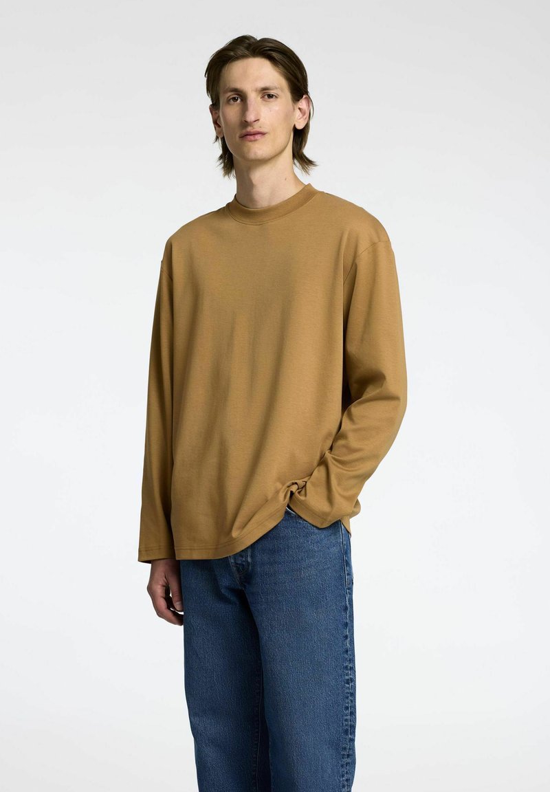 Ilgas rankovės, apvalia apykakle, marškiniai iš šilto rudo medvilnės audinio, derinami su laisvais mėlynais džinsais. Paprastas dizainas be matomų raštų ar logotipų.