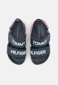 Námořnické modré sandály se dvěma širokými pásky s bílým textem "TOMMY" a "HILFIGER". Texturovaná gumová podrážka a červený, bílý a modrý lem na patě.