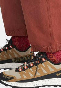 Boty na trekking vyrobené z GORE-TEXu, s béžovým a černým svrškem, oranžovými akcenty a bílými skvrnitými podrážkami, doplněné červenými vzorovanými ponožkami a volnými oranžovými kalhotami.