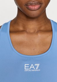 Αθλητικό τοπ σε ανοιχτό γαλάζιο χρώμα με στρογγυλή λαιμόκοψη. Διαθέτει λευκό λογότυπο "EA7 EMPORIO ARMANI" στο κέντρο. Ομαλή, ελαστική υφή υφάσματος.