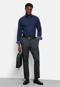 Marineblaue Hemdjacke mit gemusterten Manschetten, graue Hose, schwarzer Gürtel und Schuhe. Hält eine schwarze Aktentasche, steht vor hellem Hintergrund.