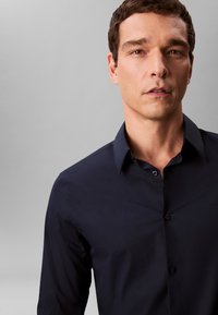 Mann mit kurzem braunem Haar, der ein dunkelblaues Hemd mit Knöpfen vor einem einfarbigen grauen Hintergrund trägt.