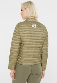 Olivgrüne Puffjacke mit gestepptem Design und Stehkragen. Verfügt über ein Etikett auf der Rückseite und wird mit passenden Hosen kombiniert.