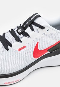 Bijela tenisica s strukturiranim mrežastim gornjim dijelom, crnim vezicama i crvenim Nikeovim znakom. Ima izražen gumeni potplat i crni akcent na peti.