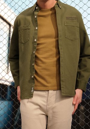 Man wearing an olive green button-up shirt over a mustard yellow t-shirt and light beige pants, standing in front of a chain-link fence.