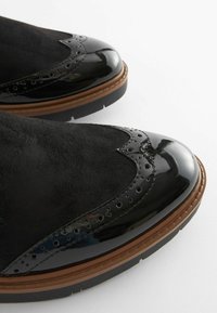 Scarpe in suede nero e pelle verniciata con dettagli brogue, dotate di una punta arrotondata e una suola marrone e nera. Superficie testurizzata.