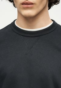 Κοντινό πλάνο ενός ατόμου που φοράει μια μαύρη φούτερ με στρογγυλή λαιμόκοψη πάνω από μια λευκή μπλούζα με στρογγυλή λαιμόκοψη, δείχνοντας το κάτω μέρος του προσώπου και τον λαιμό.