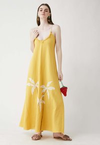 Gelbes Maxikleid mit Spaghettiträgern, fransigem Saum und weißem Blattmuster. Das Model hält eine rote Handtasche in Form einer Erdbeere. Flache Sandalen.