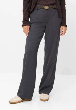 Kvinde iført mørkegrå vidde bukser med bælte, cremefarvede sneakers og en mørkebrun cardigan over en brun top, stående mod en hvid baggrund.