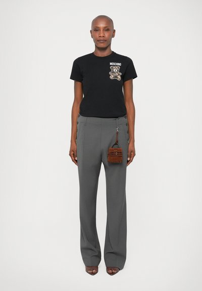 Asmuo, vilkintis juodą Moschino marškinėlį su meškiuko atspaudu, pilkas kelnes su aukštu liemeniu ir šoninėmis sagutėmis, rudą mini rankinę ir atviras bato kojines.