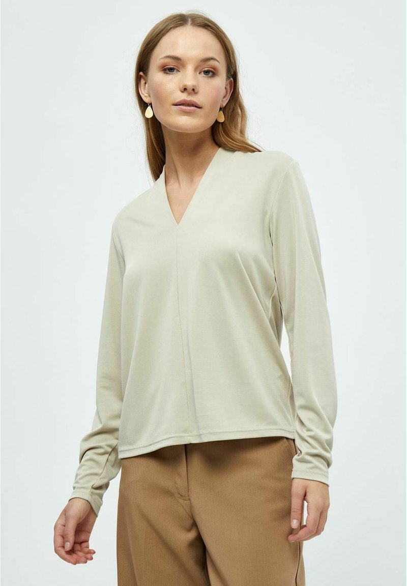 Hellbeige Langarmshirt mit V-Ausschnitt, aus glattem Stoff, mit lockerem Schnitt und subtilen Nahtdetails. Kombiniert mit braunen Hosen.