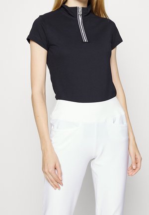 Vrouw draagt een zwart kortärmelig shirt met een ritssluiting aan de voorkant en witte hoge taillebroeken tegen een effen achtergrond.