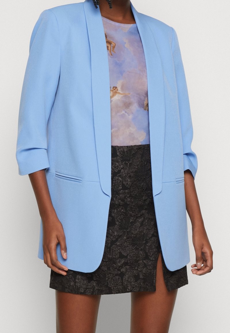 Personne portant un blazer bleu clair par-dessus un haut imprimé de nuages et une jupe noire texturée avec une fente à l’avant, sur un fond uni.