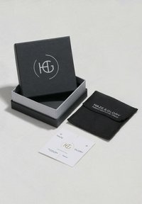 Schwarze Schmucksbox mit silbernem Logo; beinhaltet einen schwarzen Stoffbeutel und eine weiße Karte mit Markendetails. Kompakte, elegante Gestaltung.