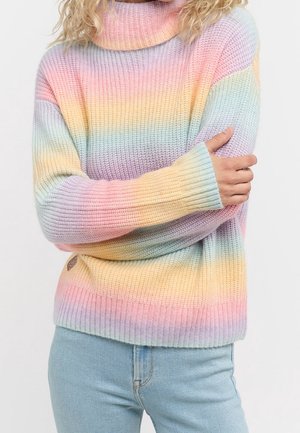 Person with curly blonde hair wearing pastel rainbow striped knit sweater with long sleeves crossed over light blue jeans.