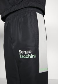 Svarta träningbyxor med en strukturerad yta, med en vit sidopanel och gröna detaljer. "Sergio Tacchini"-logotypen visas i vitt och grönt.