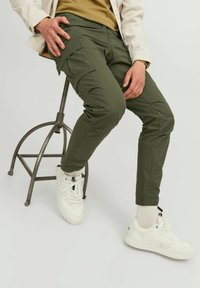 Pantaloni cargo verzi cu buzunare laterale, design slim-fit, asortați cu teniși albi și șosete cu strangere. Ales pe un scaun metalic.