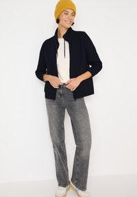 Marinblå zip-up jacka med hög krage, krämfärgad tröja under, grå jeans och en ljusgul stickad mössa. Avslappnade skor med accenter.