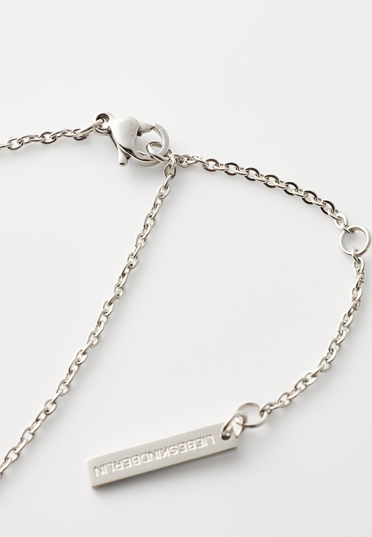 Silberne Kettenarmband mit einem Karabinerhaken, das ein rechteckiges Etikett mit geprägter Schrift trägt. Die Kettenglieder sind klein und fein strukturiert.