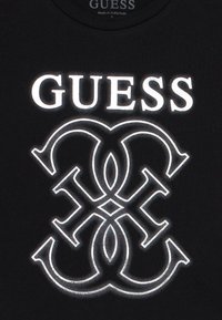 Μαύρο βαμβακερό t-shirt με μεγάλο λευκό λογότυπο "GUESS" πάνω από έναν περίπλοκο διαπλεκόμενο σχεδιασμό σε λευκό, με λεία υφή.