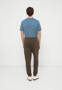 Mann steht mit dem Gesicht zu einer weißen Wand, trägt ein blaues kurzärmeliges Hemd, olivfarbene Jogginghosen und weiße Turnschuhe mit Gummisohlen.