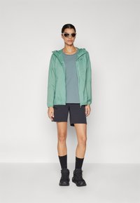 Light green hooded jacket over a teal shirt, paired with black shorts and black hiking boots. White sunglasses complete the outfit.