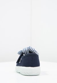 Zapato de lona azul marino con suela de goma blanca, que presenta un lazo a rayas azul y blanco. La vista trasera muestra un tejido texturizado y un diseño simple.