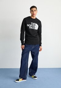 Sudadera negra con el logo blanco de "THE NORTH FACE", combinada con unos vaqueros de mezclilla azules holgados. Modelo con zapatillas negras y amarillas. Fondo sencillo.