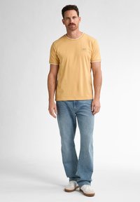 Tricou galben cu mânecă scurtă și margini albe, asociat cu blugi albaștri deschiși și adidași albi. Materialul pare neted și are un design casual.