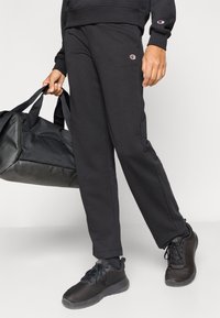 Μαύροι φόρμες από μαλακό ύφασμα, με λογότυπο στο αριστερό μηρό, συνδυασμένοι με μαύρα αθλητικά παπούτσια και μαύρη τσάντα γυμναστηρίου.